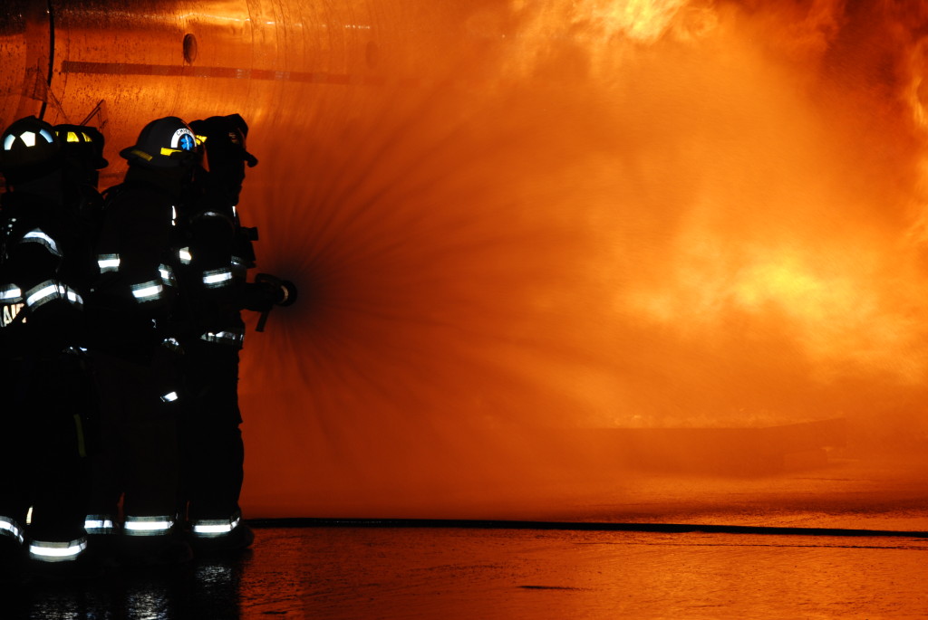 Action photography, Four firefighters in black uniforms with bright reflective stripes spraying water on a large fire.