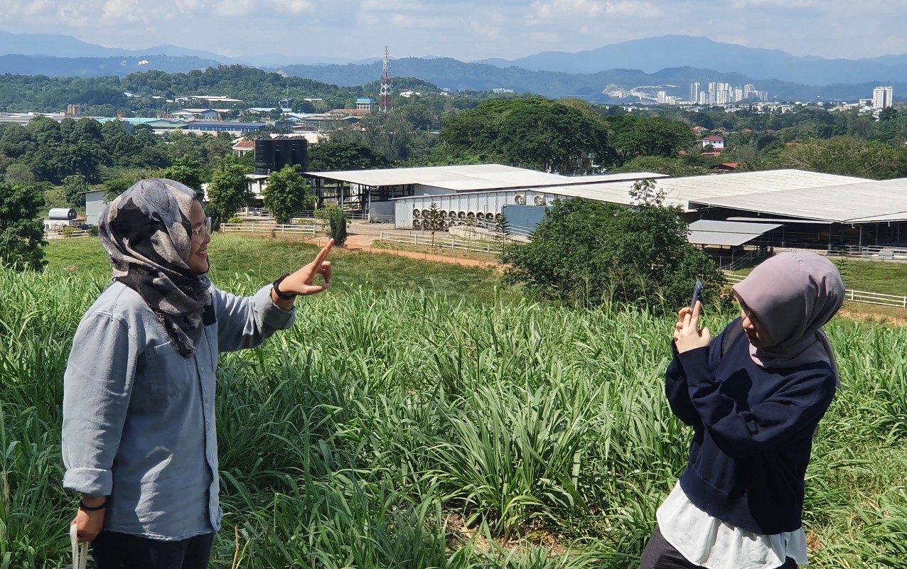 Two women wearing hijabs taking a picture of each other on a grass-covered hill overlooking a dairy farm.
