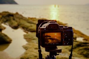 An image of a camera mounted on a tripod capturing a photograph of a sunset from the shore